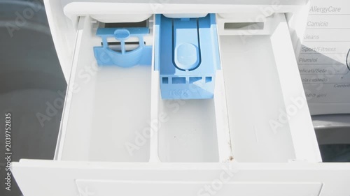 Pouring liquid laundry detergent into washing machine dispenser drawer from above. Close-up overhead view shows blue compartments and hand holding bottle. Domestic cleaning.
