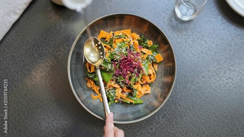 A vibrant and artfully presented noodle dish, showcasing broad, flavorful noodles tossed with fresh green vegetables and garnished with a sprinkle of white and black sesame seeds and delicate red