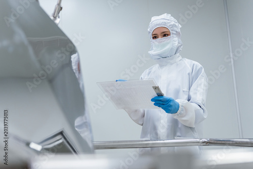 She works near mixing equipment in a high-containment lab. Professional scientists in cleanroom suits inspecting pharmaceutical manufacturing processes and operating industrial mixing equipment.
