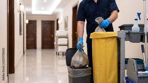 Professional hotel housekeeper wearing blue gloves collecting trash bags from a hallway bin into a janitorial cleaning cart trolley in a luxury resort corridor