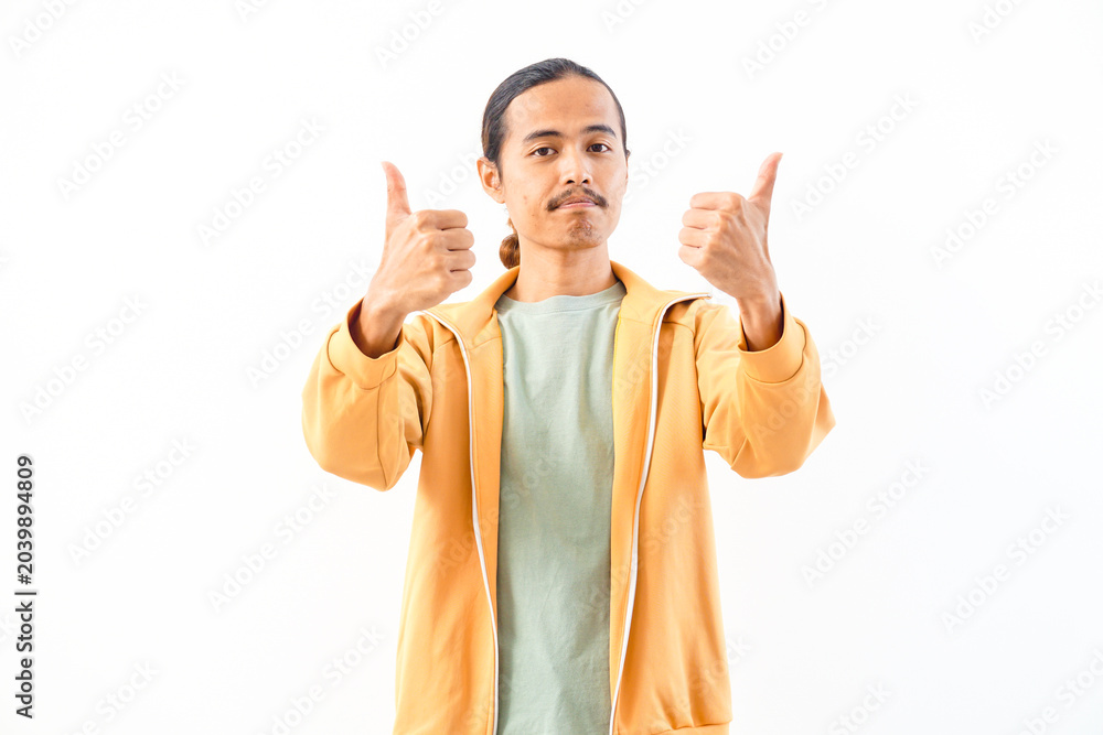 Naklejka premium Young Asian male wearing a beige jacket smiles while making a double thumbs-up gesture indoors.