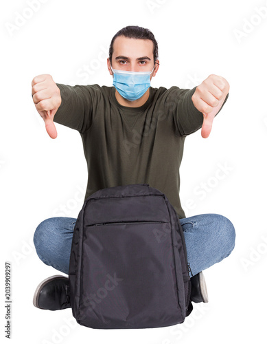 Картината върху платно Seated discontent man traveler with a backpack, wearing face mask as protection against different virus, shows thumbs down negative gesture isolated on transparent background