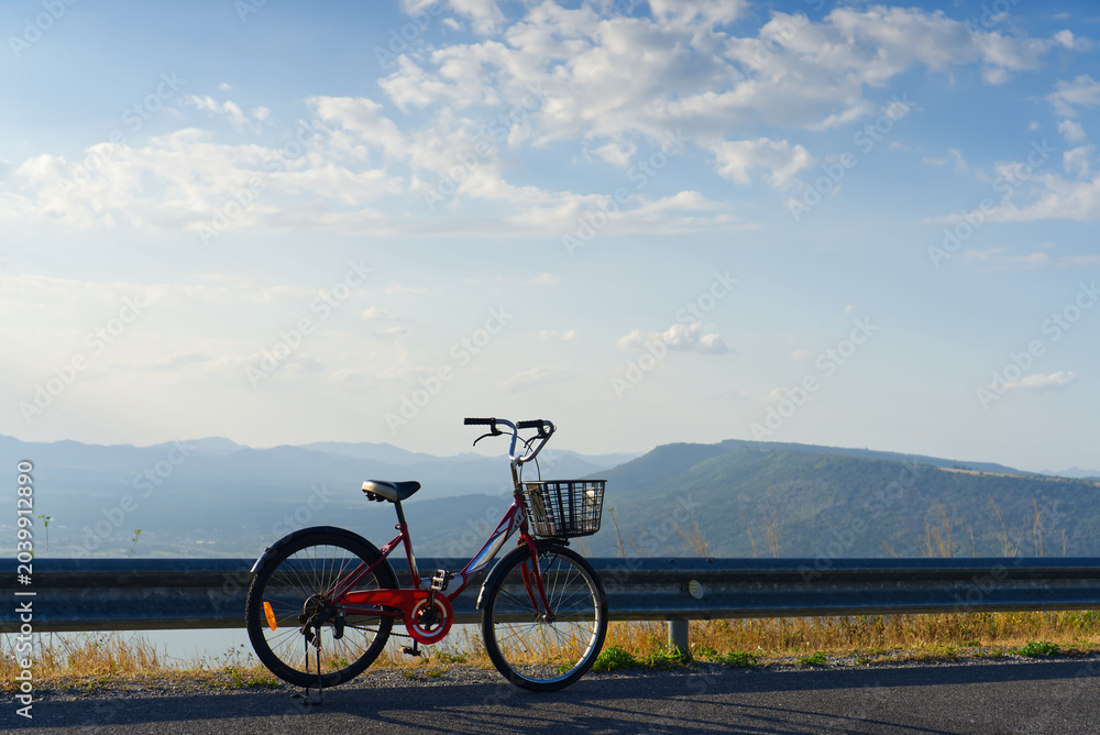 Fototapeta premium Red bicycle parked on the roadside with a beautiful scenic mountain landscape under a bright blue sky.