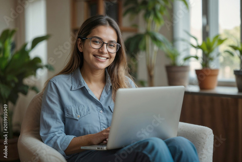 Portrait of happy European woman with glasses uses laptop while sitting in armchair at home