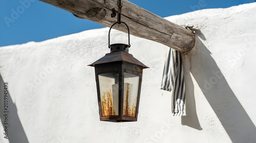 Rustic metal lantern hanging from wooden beam against white wall and blue sky.
