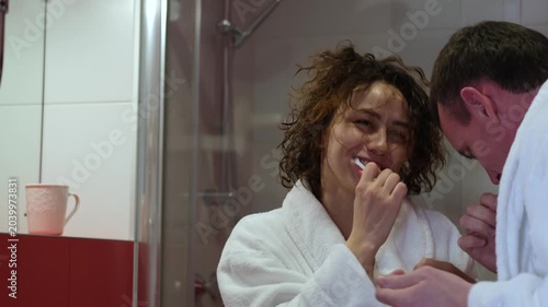 A married couple in white coats stands in the bathroom and brushes their teeth for oral hygiene. Morning routine together at home. A young man and woman with a toothbrush stand near a mirror in the