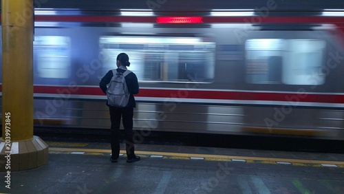 Bild auf Leinwand A lone commuter with a backpack stands on a station platform as a commuter train rushes past with dynamic motion blur