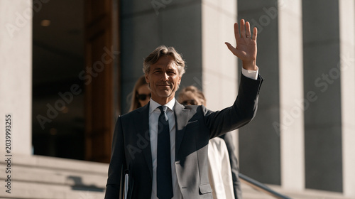 Finance minister emerging from a government building with advisors, arm raised to hold back press, budget folders tucked under arm, July morning light harsh on concrete steps, breaking news fiscal a
