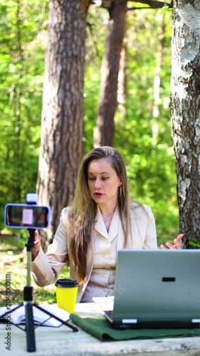Online work away from the confines of a stuffy office in a bustling city. A beautiful 40-year-old woman conducts an online consultation in front of a smartphone camera in a city park or forest.
