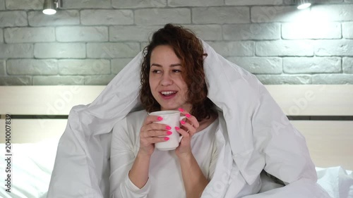Young beautiful woman drinks water from a mug while sitting in bed in a cozy bedroom. Morning rituals.