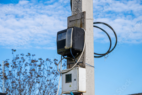 Fotografie An old black electrical meter and a white metal junction box are mounted on a wooden board attached to a concrete utility pole under a partly cloudy blue sky