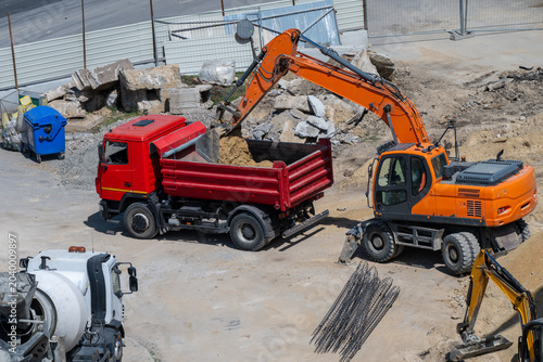 a bright orange wheeled excavator loading dirt and rubble into a red dump truck on an active, sunlit construction site.