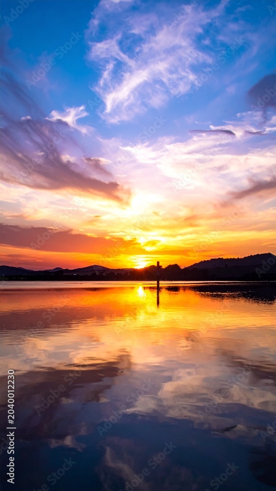 Fototapeta premium A breathtaking vertical view of a vibrant sunset over a calm lake with silhouetted mountains reflecting in the peaceful water surface at dusk.