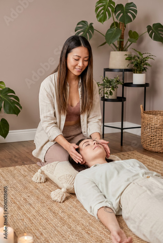 Asian woman practitioner performing reiki energy healing therapy or head massage on a relaxed female client in a cozy wellness studio.
