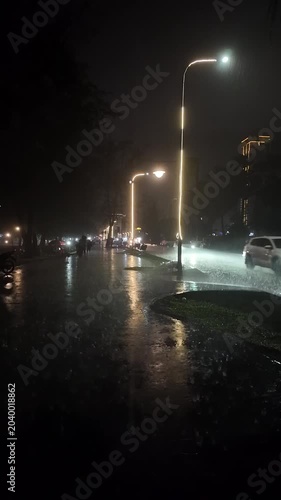 Vertical night shot of heavy rain pouring on a city street, where cars, motorbikes, and vehicles circulate under glowing street lamps, reflecting on the wet, shimmering pavement.