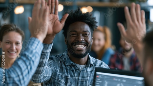 Candid shot of a diverse team celebrating a project milestone with high-fives and smiles around a computer monitor showing good results, joyful, successful, authentic teamwork