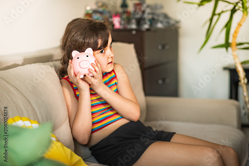 A young girl in a striped tank top sitting on a couch, putting a coin into a pink piggy bank