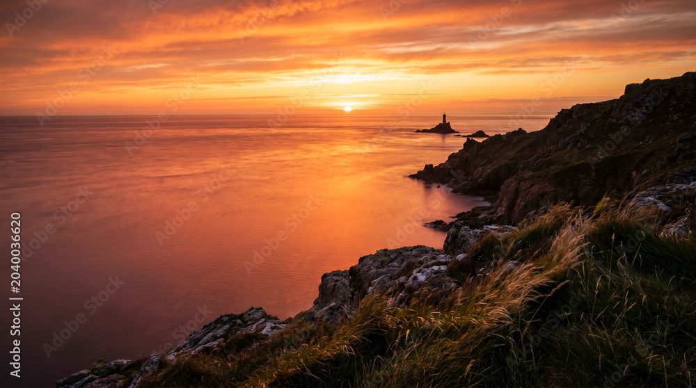 Obraz premium Puesta de sol desde un acantilado costero sobre un mar en calma en verano, con el agua pintada de naranja y rojo y larga exposición suavizando las olas.