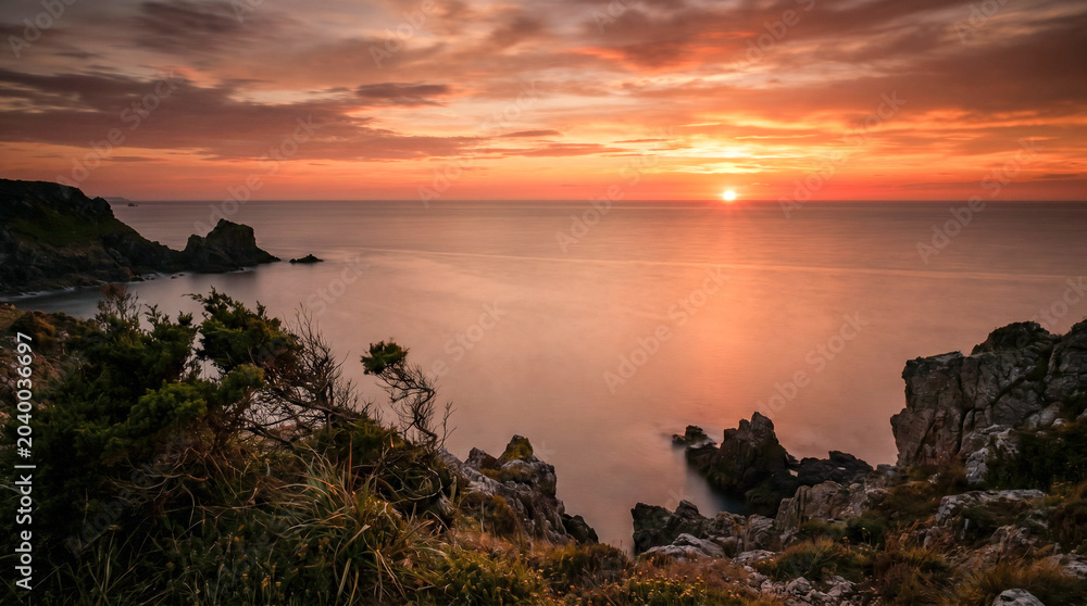 Fototapeta premium Puesta de sol desde un acantilado costero sobre un mar en calma en verano, con el agua pintada de naranja y rojo y larga exposición suavizando las olas.