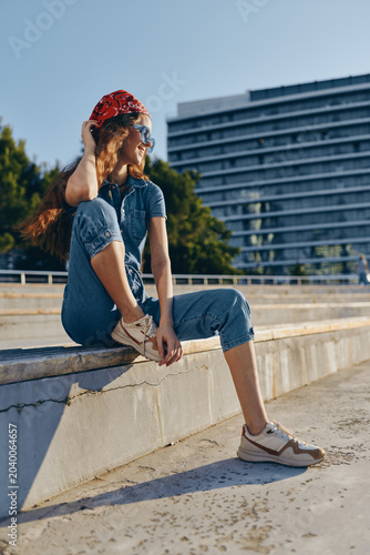 Fotografía young woman sitting outdoors in casual denim outfit with red bandana and sunglas