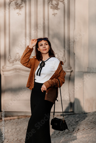 Beautiful young woman in sunglasses going for a walk in Prague city. Stylish girl in a brown jacket posing near a historic building facade on a sunny day. Urban fashion and lifestyle concept.