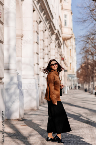 Beautiful young woman in sunglasses going for a walk in Prague city. Stylish girl in a brown jacket posing near a historic building facade on a sunny day. Urban fashion and lifestyle concept.