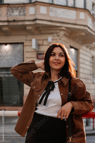 Beautiful young woman in sunglasses going for a walk in Prague city. Stylish girl in a brown jacket posing near a historic building facade on a sunny day. Urban fashion and lifestyle concept.