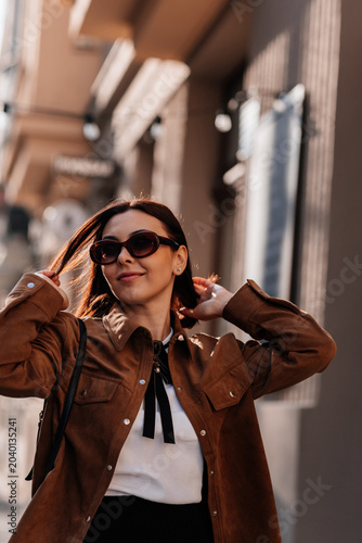 Beautiful young woman in sunglasses going for a walk in Prague city. Stylish girl in a brown jacket posing near a historic building facade on a sunny day. Urban fashion and lifestyle concept.