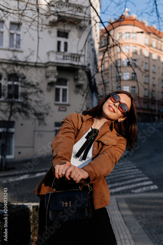 Beautiful young woman with a handbag standing near historic building in Prague. Stylish girl in sunglasses and a fashionable outfit enjoying a sunny day in city. Urban fashion and lifestyle concept.