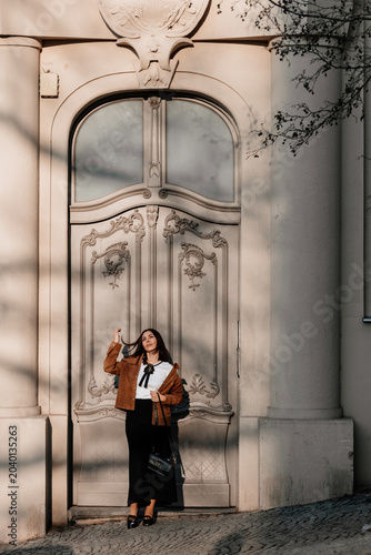 Beautiful young woman in sunglasses going for a walk in Prague city. Stylish girl in a brown jacket posing near a historic building facade on a sunny day. Urban fashion and lifestyle concept.