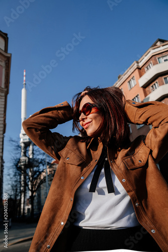 Beautiful young woman in sunglasses having fun in Prague city. Stylish girl in trendy brown jacket posing on urban street with TV tower on sunny day. Fashion portrait at Zizkov, trendy outfit