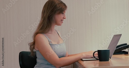 Young woman typing on laptop at desk in office, then leaning back on chair to rest and taking black coffee mug. Medium side profile shot of remote worker or student during daytime. Slow motion.