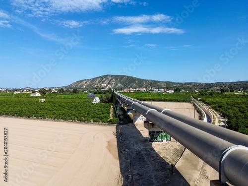 Aerial view of the large Tajo Segura water transfer pipeline crossing the fertile agricultural lands orchards and fields of Vega Baja under a clear blue sky towards a distant mountain