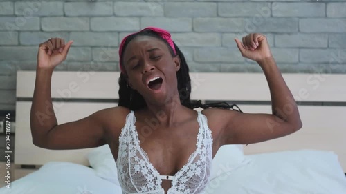 A very beautiful dark-skinned woman wakes up, takes off her sleep mask, stretches her arms and yawns in the bedroom.
