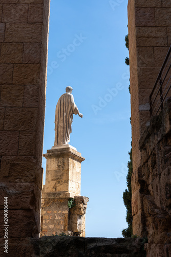 Valokuva A majestic stone statue of a classical Roman figure viewed from behind, standing tall against a clear blue sky and dramatically framed by two towering ancient ruins