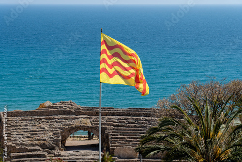 Canvas Print The official flag of Catalonia blowing in the wind against a vibrant blue seascape, with the historic stone archways of Tarragona's Roman amphitheater below