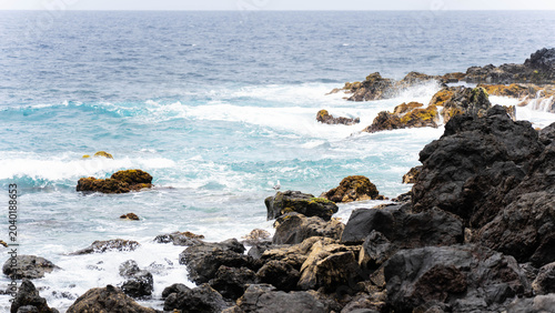 Fotografie Close up of powerful turquoise ocean water and white sea foam crashing against black volcanic rocks along the rugged shoreline