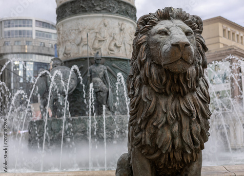 bronze lion statue guarding a grand fountain with water sprays and classical historical monument in the background city center square