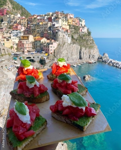 Delicious gourmet bruschetta served above a picturesque Mediterranean coastal village. Fresh ingredients, vibrant colors, and stunning sea views create an irresistible travel and food experience.