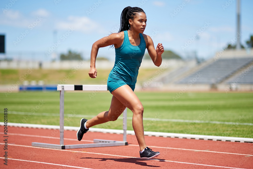 Obraz premium Athletic woman running and clearing hurdle on track field