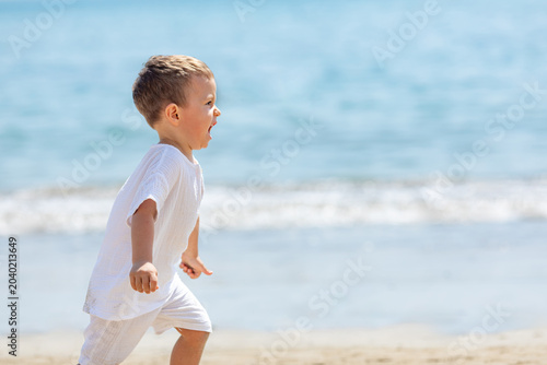 Fotografie Happy toddler child boy in white outfit joyfully runs along the sandy beach with the ocean waves gently lapping in the background