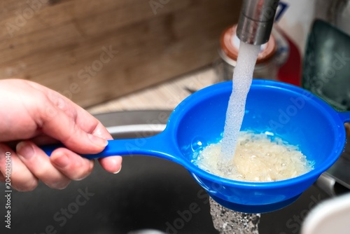 Washing a small amount of white rice in a blue plastic sieve under running kitchen tap water