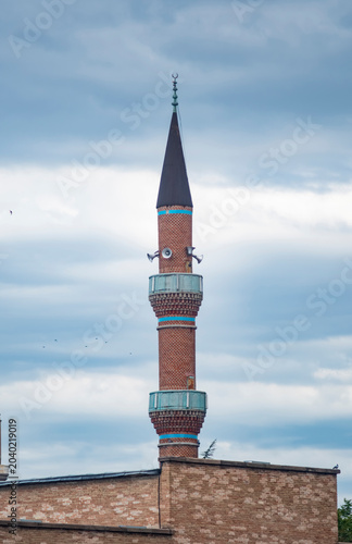 Traditional brick minaret tower against cloudy sky. A historic red brick minaret with loudspeaker horns stands tall under a dramatic overcast sky. Showcasing traditional Islamic architecture.