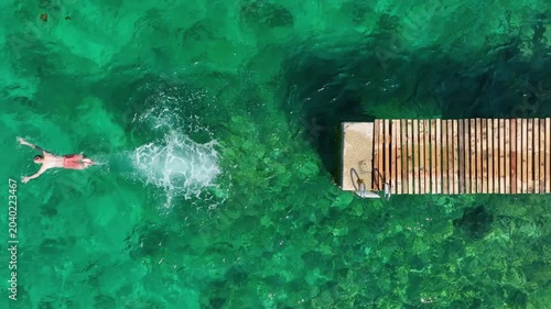 Aerial top view of a person jumping from a wooden pier into the clear turquoise sea water. Scenic drone video of a traveler enjoying a summer vacation trip.
