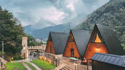 Wooden A-frame cabins with black metal roofs surrounded by green mountains and misty clouds. Concept of eco-tourism, glamping, nature escape, and cozy sustainable travel