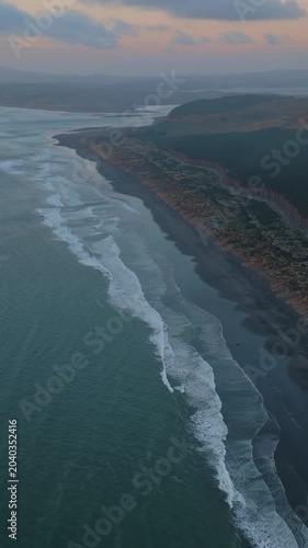 Aerial view of the coastline at sunset, showing the beach, ocean waves, and forest in Kawhia, New Zealand. The scene captures the natural beauty of the landscape.