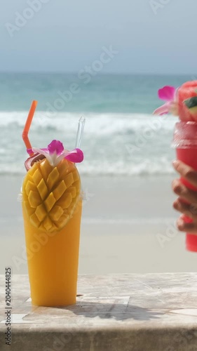 Vertical video. Young woman reaching for and drinking a watermelon smoothie from a table where a mango smoothie also sits, with the sunny beach and sea behind her. Tropical refreshment concept