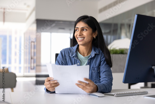 Canvastavla Indian woman sits at desk in office, holding documents, smiling, looking away, f