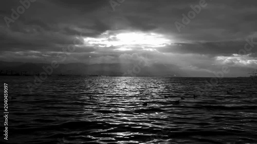 Dark grey cloudy sky Gloomy sky over rippling sea with birds, light rays beaming onto distant mountains. Ideal for background, nature or atmospheric concept.