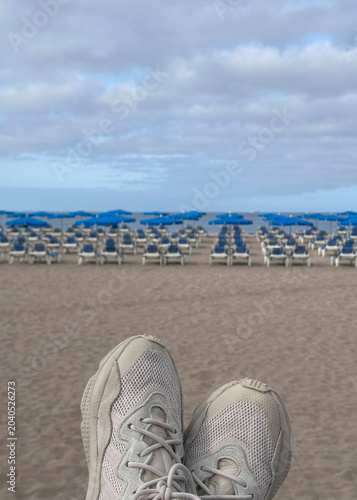 Pair of sneakers with a blurred beach scene in the background,rows of blue umbrellas a ocean.perfect a reel about a beach vacation,   relaxation, or an adventure to the sea.